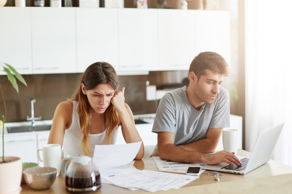 young caucasian couple having concentrated look, doing paper work and working with laptop computer while sitting in kitchen. young family couple managing finances, checking their family budget